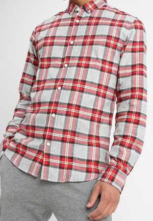 Man wearing a red, gray, and black plaid button-up shirt with long sleeves and gray pants, standing with one hand partially visible.
