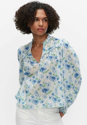 Kvinde med krøllet hår iført en gennemsigtig hvid bluse med blåt blomsterprint og puffede lange ærmer, kombineret med hvide bukser.