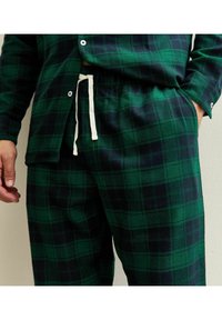 Pigiama di flanella a quadri verdi e blu scuro, composto da una camicia con bottoni e una fascia elastica in vita, con un laccio bianco e tessuto testurizzato.