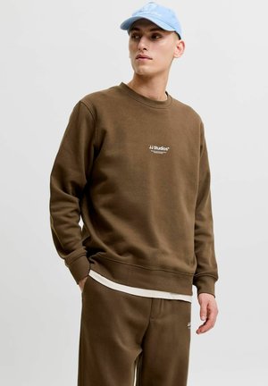 Jeune homme portant un sweat-shirt marron JJ Studios et un pantalon assorti avec une casquette de baseball bleu clair, debout sur un fond gris uni.