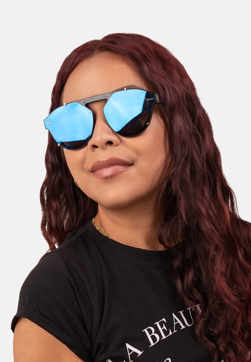 Black octagonal sunglasses with blue mirrored lenses, metal accents, and a thick frame, worn by a person with wavy, dark red hair.