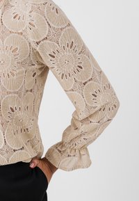 Lace blouse in beige met bloemenpatronen. Heeft lange mouwen met gerimpelde manchetten en een semi-doorzichtige textuur.