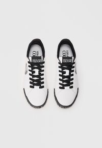 Balti sportiniai batai su tekstūruotu paviršiumi, juodomis raištelėmis, juodais akcentais ant apykaklės ir logotipo etikete ant liežuvio. Guminis padas.