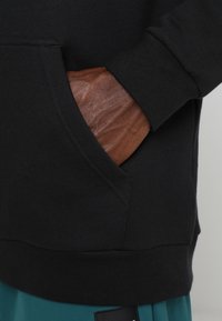 Mano con pelle scura appoggiata all'interno della tasca frontale di una felpa nera con pantaloni blu-verde visibili sotto.