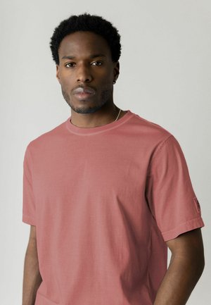 Mann med kort krøllete hår iført en rosa t-skjorte og sølvkjede, stående foran en ensfarget lys grå bakgrunn.