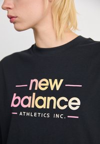 Šviesiaplaukis asmuo dėvi juodą marškinėlį su spalvingu "new balance ATHLETICS INC." logotipo, matomas nuo kaklo iki krūtinės vidurio, pasuktas į kairę.