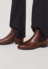 Stivaletti in pelle marrone alla caviglia con pannelli elastici ai lati e un tacco basso a blocco, abbinati a pantaloni neri, che mostrano una finitura liscia e lucida.