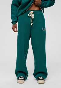 Teal sweatpants in een ontspannen pasvorm met een trekkoord in de taille, zijzakken en geborduurd logo-detail. Het materiaal lijkt zacht en gestructureerd.