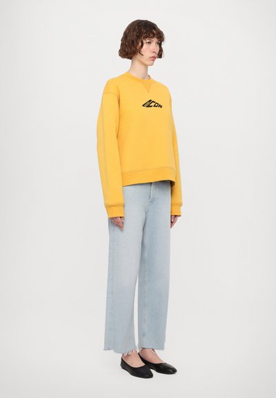 Gul crew-neck sweatshirt med svart logo, kombinert med lysblå vide jeans med frayed hem og svarte leiligheter.