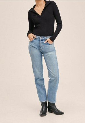 Kobieta ubrana w czarną bluzkę z długim rękawem, jasnoniebieskie wysokie jeansy i czarne botki, z rękami w kieszeniach.