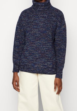 Personne portant un pull à col roulé en maille mêlée bleu marine et blanc et un pantalon large écru sur un fond uni.