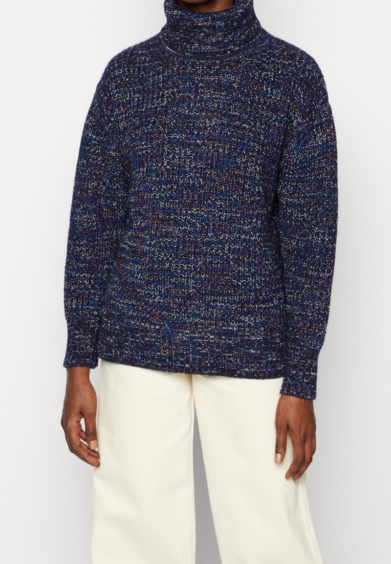 Personne portant un pull à col roulé en maille mêlée bleu marine et blanc et un pantalon large écru sur un fond uni.