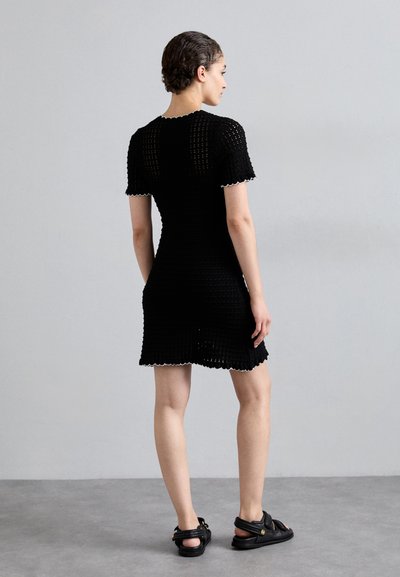 Schwarzes gestricktes Kleid mit kurzen Ärmeln, gewellten Rändern und taillierter Passform. Trägt man mit schwarzen Sandalen, von hinten betrachtet.