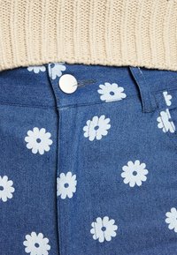 Jeans de ganga com um padrão floral azul, apresentando margaridas brancas, um fecho de botão frontal e um suéter de malha texturizada em creme por cima.