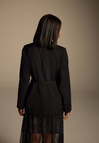 Black blazer with a fitted waist, tailored design, and long sleeves, paired with a translucent black lace skirt, showcasing detailed texture.
