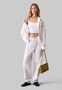 Giacca di denim bianca sopra un crop top bianco e pantaloni larghi bianchi. Il modello tiene una borsa gialla testurizzata e indossa scarpe nere.