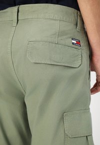 Pantaloni cargo verdi in cotone con una tasca posteriore, dettagli cuciti e un piccolo patch logo con accenti rossi, bianchi e blu.