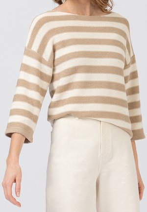 Femme portant un pull en maille beige et blanc à rayures horizontales avec des manches trois-quarts et un pantalon beige clair sur un fond uni.