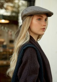 Cappello a spina di pesce grigio, lunghi capelli biondi, indossando un cappotto di lana marrone scuro con una tracolla nera texture. Sfondo sfocato.