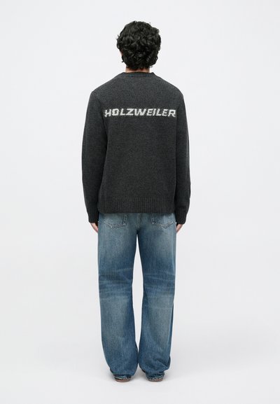 Sötétszürke pulóver kerek nyakkal, a hátán világosabb szürke "HOLZWEILER" felirattal. Laza, világoskék farmernadrággal viselve.