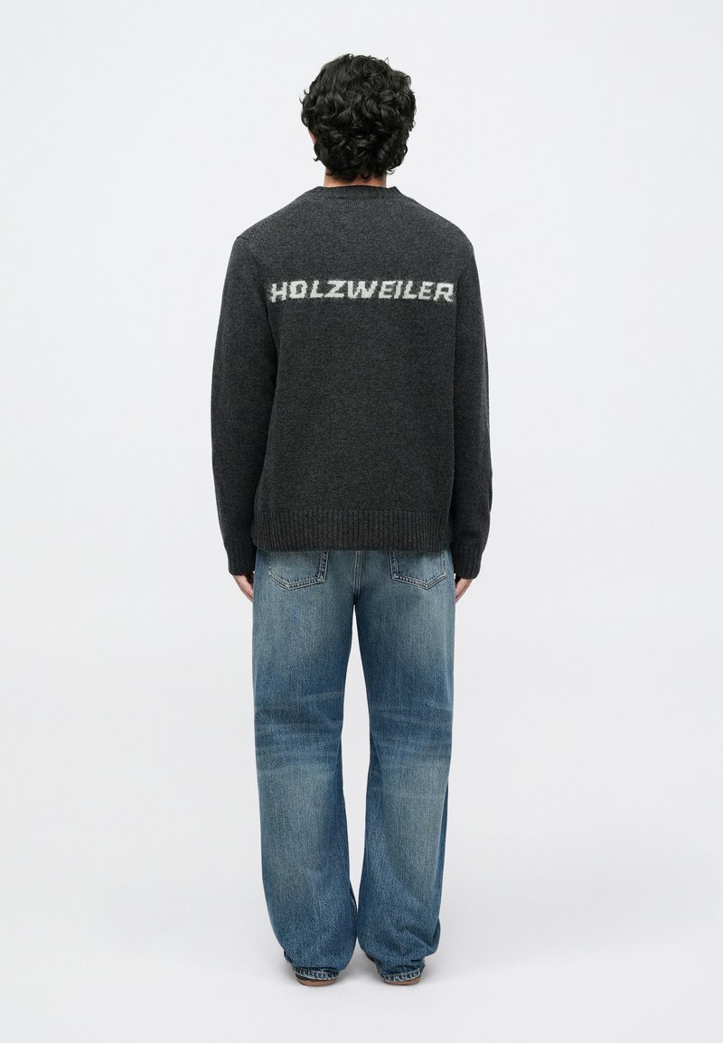 Pull gris foncé à col rond, avec "HOLZWEILER" en gris plus clair au dos. Porté avec un jean denim bleu clair ample.
