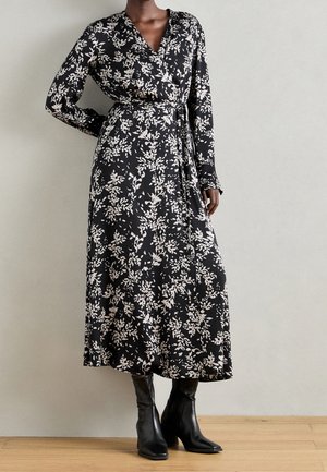Femme portant une robe portefeuille noire à motifs floraux blancs et des bottines à talons noires, debout contre un mur clair uni.