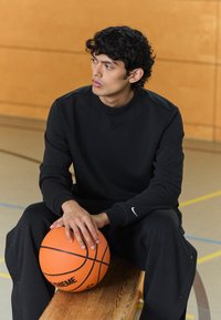 Sudadera negra y pantalones de chándal, tejido texturizado, diseño ajustado. Sosteniendo un balón de baloncesto naranja con acentos negros, sentado en un banco de madera.