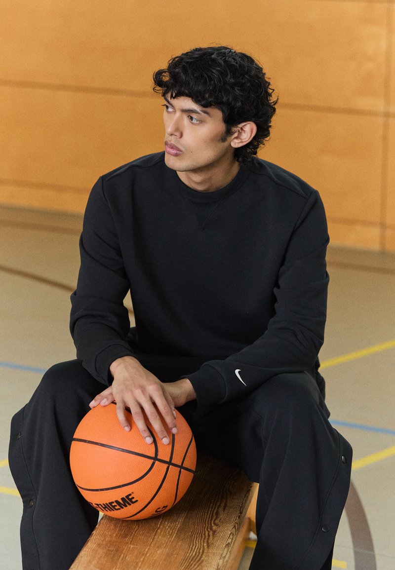 Sudadera negra y pantalones de chándal, tejido texturizado, diseño ajustado. Sosteniendo un balón de baloncesto naranja con acentos negros, sentado en un banco de madera.