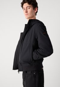 Juoda gobtuvinė striukė su siuvinėtu tekstūru, priekyje užtrauktukas, šoniniai kišeniai ir gumuotas apykaklės kraštas. Oda logotipo detalė dešiniajame rankovėje.