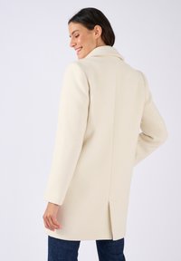 Cappotto beige chiaro con colletto a punte, vestibilità su misura e maniche lunghe. Presenta una fessura centrale sul retro e un tessuto liscio e texturizzato.