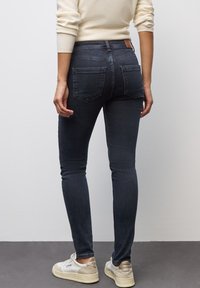 Los jeans skinny de mezclilla oscura presentan una cintura de altura media, bolsillos traseros y un parche de cuero marrón. Combinados con zapatillas blancas.