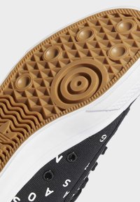 Svart canvas sneakers med vita detaljer, med en texturerad brun gummisula med cirkulära och triangulära mönster för grepp.