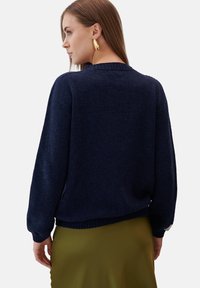 Granatowy sweter dzianinowy z długimi rękawami, ściągaczami przy mankietach i u dołu. Miękka tekstura; noszony na zielonej satynowej spódnicy. Minimalistyczny design.