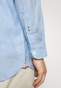 Chemise en lin bleu clair avec des poignets boutonnés, une poche avant simple et un ourlet incurvé, avec une sous-couche rayée visible.