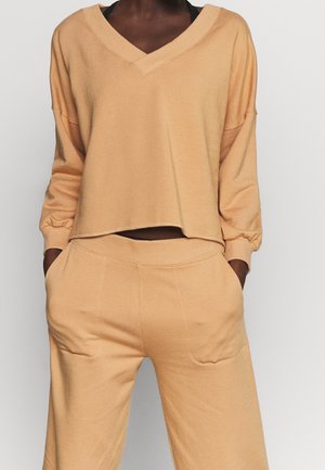 Osoba ubrana w luźną jasnobrązową bluzkę z długim rękawem i dekoltem w serek oraz dopasowane szerokie spodnie, z rękami w kieszeniach, na jednolitym jasnym tle.