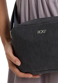 Grijze ribcord tas met zilverkleurige hardware, voorzien van een gestructureerd geribbeld ontwerp en een schouderriem, vastgehouden in een hand tegen een lichte stof achtergrond.