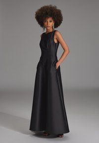 Schwarzes Satinabendkleid mit tailliertem Oberteil und weitem Bein. Mit Rundhalsausschnitt und seitlichen Taschen. Glatte Textur, minimalistisches Design.