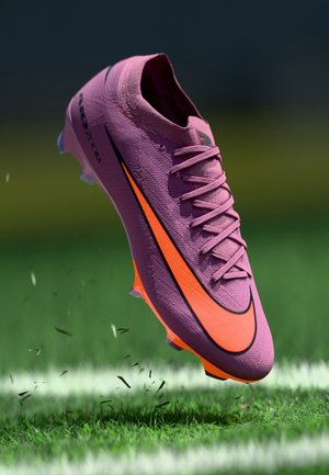 Purple and orange soccer cleat with laces suspended mid-air above grass field, showing side view and Nike swoosh logo.