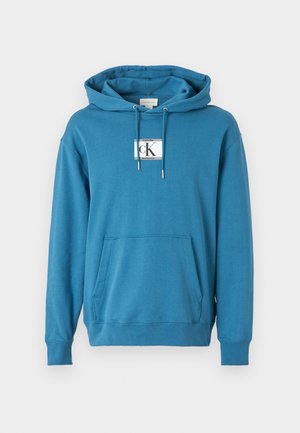 Blå hettegenser laget av myk stoff, med frontlomme, ribbede mansjetter, og en rektangulær logo-patch på brystet.