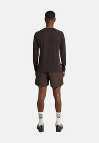 Mann, der abgewandt steht, tr�ägt ein langärmliges dunkles Hemd, dunkle Shorts, weiße Socken mit dem Text "OFF DUTY GEAR" und schwarze und beige Sneaker.