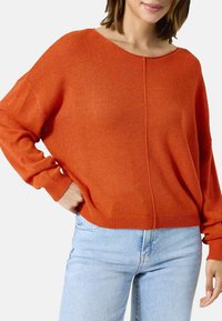 Oranje gebreide trui met een ontspannen pasvorm, brede halslijn en lange mouwen. De zoom is geribbeld, wat zorgt voor een gestructureerde afwerking. Gecombineerd met lichte jeans.