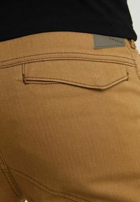 Zbliżenie brązowej teksturowanej kieszeni spodni z klapką i szarym logo marki, noszonej z czarnym topem.