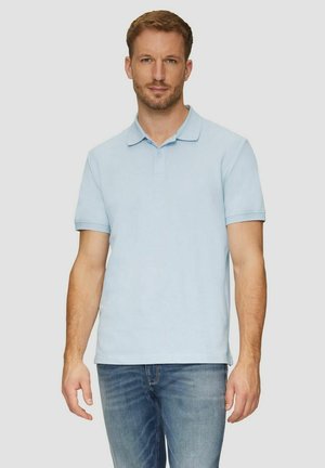 Mann in hellblauem kurzärmligem Poloshirt und blauer Jeans steht vor einem einfarbigen hellen Hintergrund und blickt nach vorne.