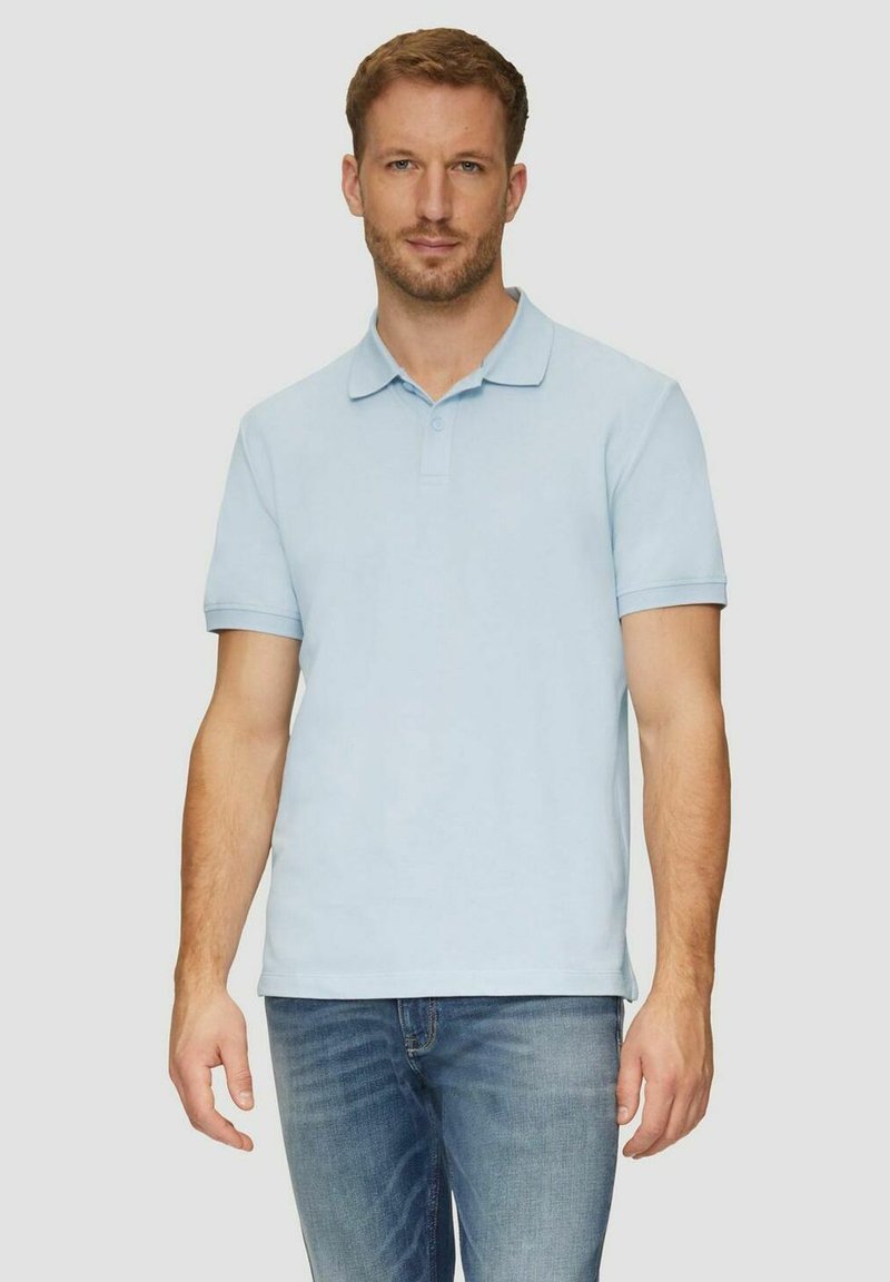 Mann in hellblauem kurzärmligem Poloshirt und blauer Jeans steht vor einem einfarbigen hellen Hintergrund und blickt nach vorne.