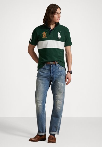Zaļš polo krekls ar baltu horizontālu svītru, izšuvēts logo, apvienots ar nolietotām zils džinsiem un brūnām ādas kurpēm.