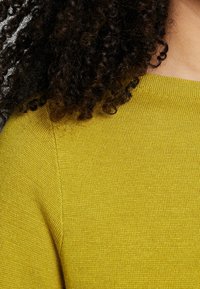 Cheveux bouclés et foncés reposant sur l'épaule, portant un pull en tricot jaune moutarde avec un col rond, montré en gros plan.