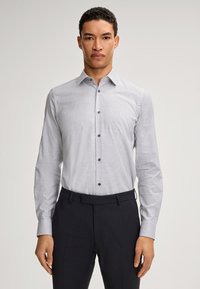 Helle graue Button-Up-Bluse mit langen Ärmeln, subtilen Karomuster, dunklen Knöpfen, kombiniert mit dunklen Hosen, die eine maßgeschneiderte Passform zeigen.