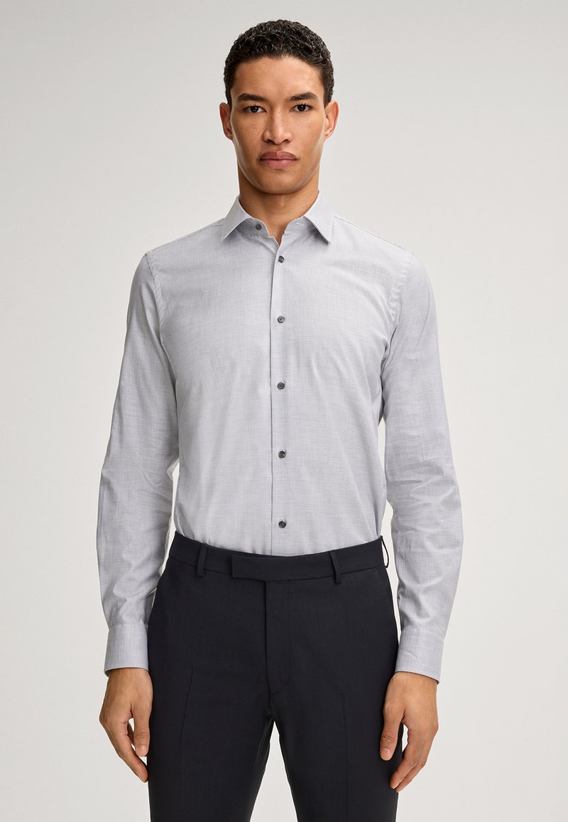 Helle graue Button-Up-Bluse mit langen Ärmeln, subtilen Karomuster, dunklen Knöpfen, kombiniert mit dunklen Hosen, die eine maßgeschneiderte Passform zeigen.