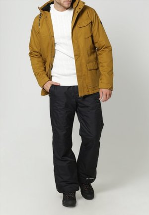 Homme portant une veste jaune moutarde, un pull en maille torsadée blanc, un pantalon de neige noir et des chaussures de randonnée noires, debout les mains dans les poches.