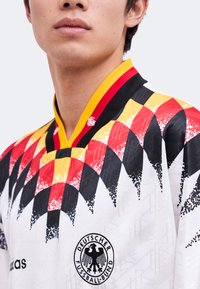 Nahaufnahme einer Person, die ein weißes deutsches Fußballtrikot mit rot-schwarz-gelben karierten Mustern und dem Logo des Deutschen Fußball-Bundes trägt.